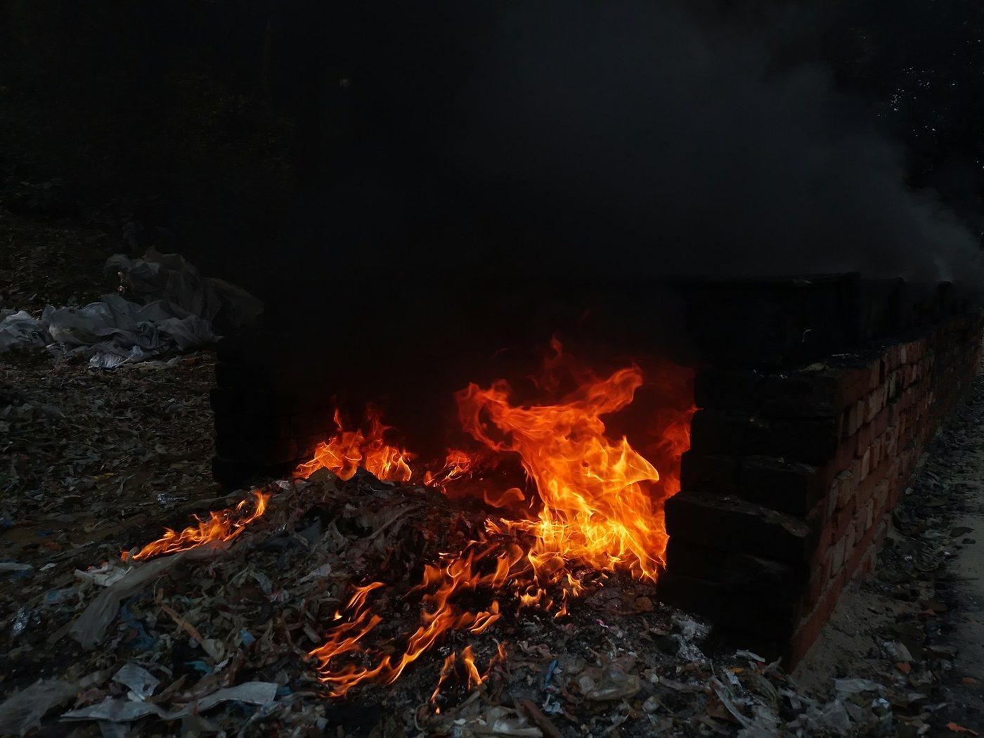 A heap of garbage burning beside a wall, dark smoke rising